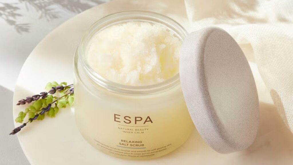 ESPA Relaxing Salt Scrub jar opened to show its grainy texture, placed beside herbs, known for gently exfoliating and softening skin with a spa-like feel.