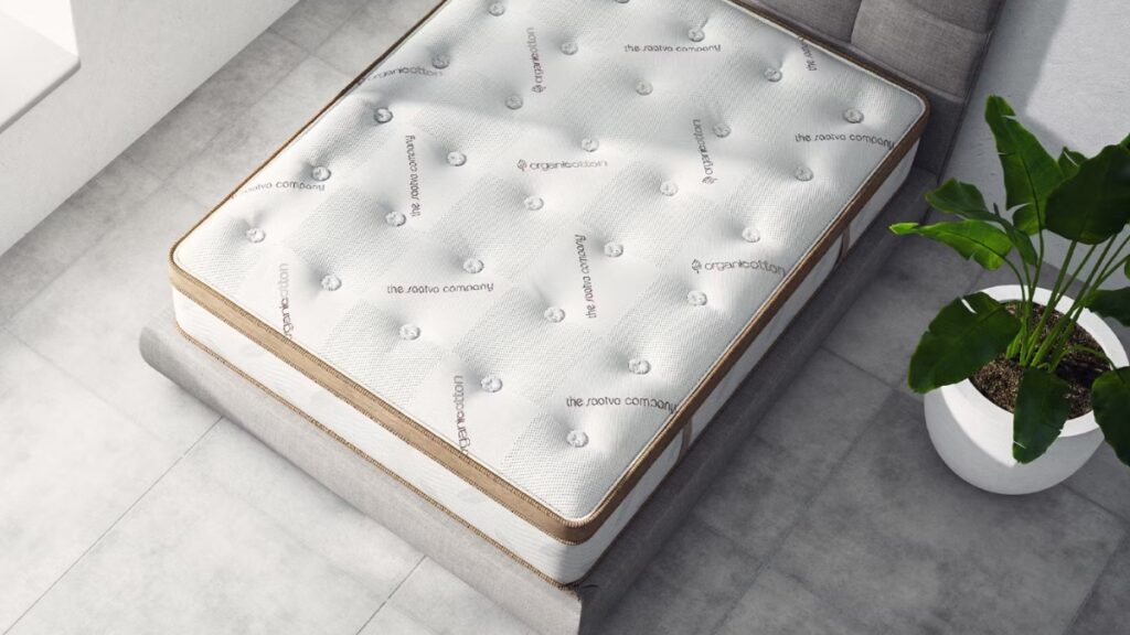 Saatva mattress with an organic cotton cover on a light grey bed frame in a bright room with tiled flooring and a potted plant.