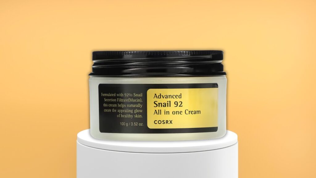 COSRX Advanced Snail 92 All in One Cream in a 100g jar shown on a white stand with a yellow background, a popular pick among best skincare products.