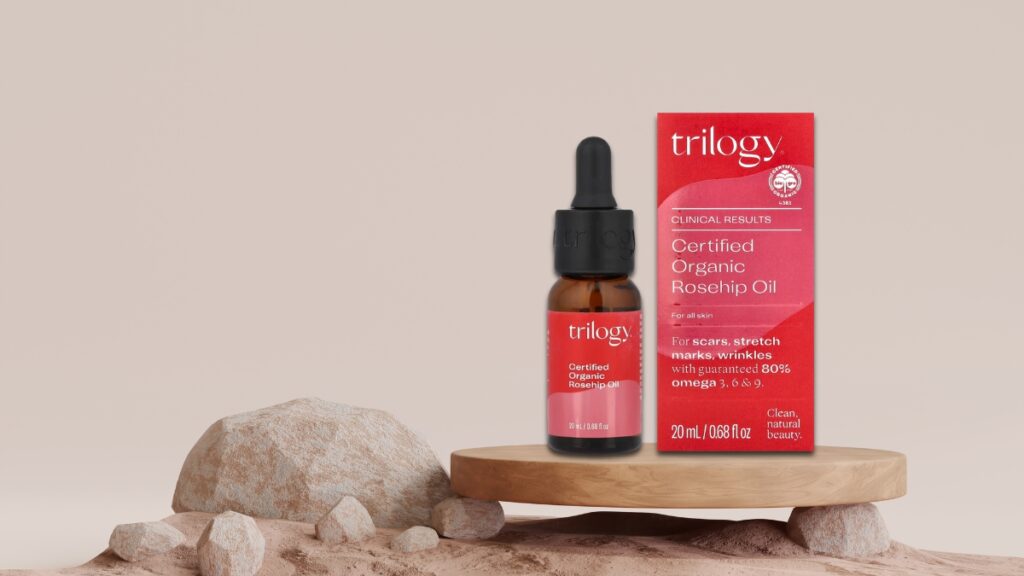 Trilogy Certified Organic Rosehip Oil in a 20ml dropper bottle with box packaging, shown on a wooden stand with rocks, a trusted skincare product for scars and fine lines.