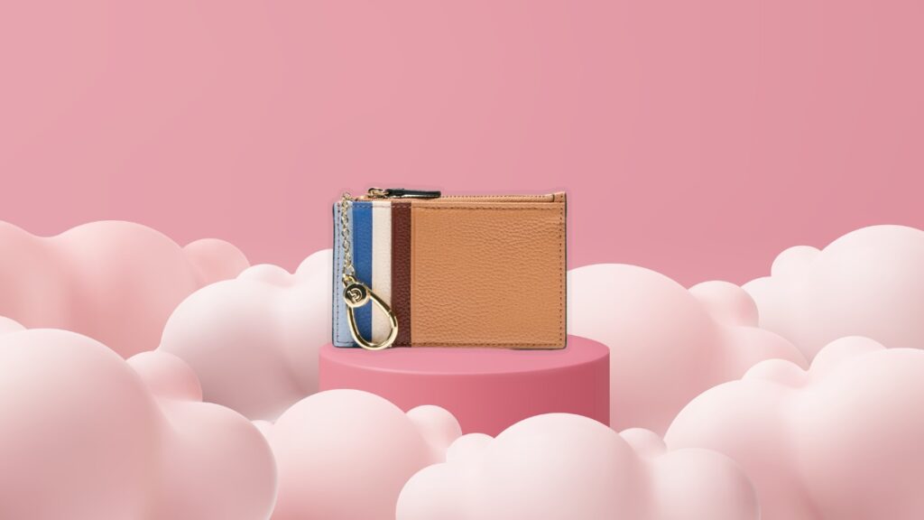 A multicolor Cole Haan card case with gold keychain detail displayed on a pink pedestal surrounded by soft cloud shapes.