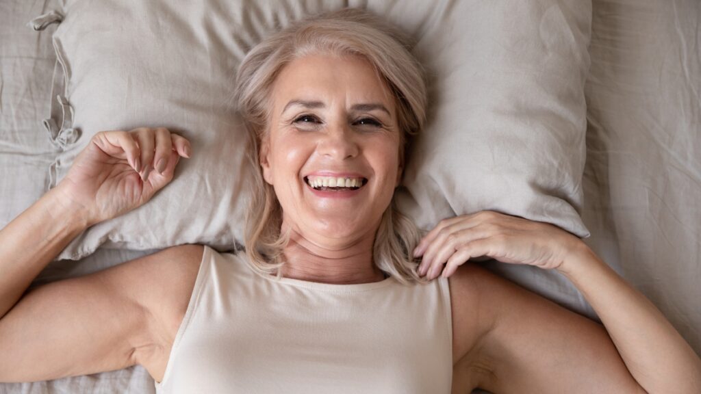 Smiling older woman with gray hair lying on a pillow, looking happy and relaxed.